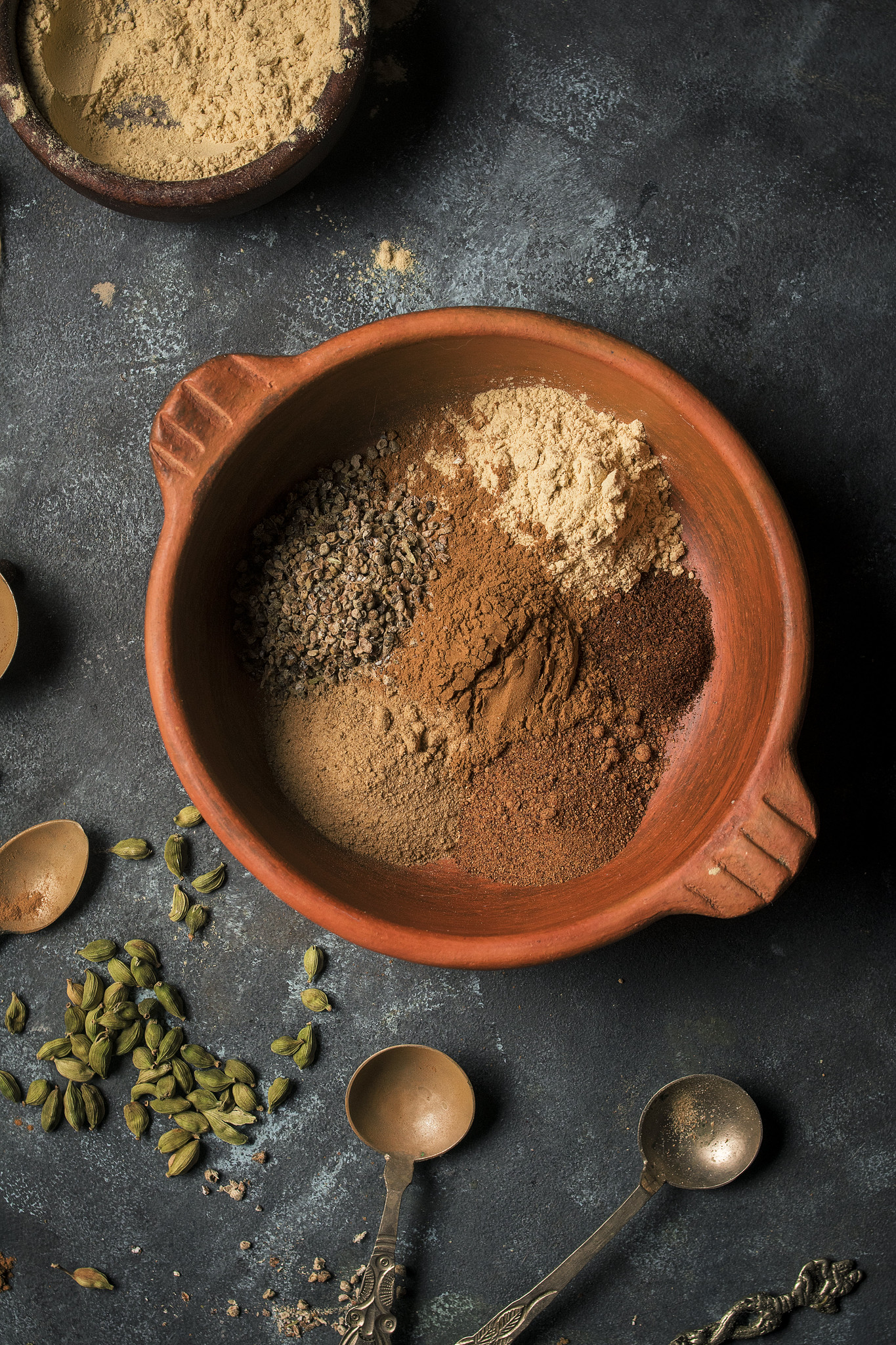 Ground spices measured into a terracotta bowl with cardamom pods and measuring spoons.